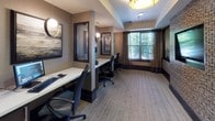 This single-floor business center is thoughtfully designed to provide a functional and refined workspace. The room accommodates multiple individual workstations, each equipped with a computer, keyboard, and ergonomic task chair, making it well-suited for guests or professionals needing a dedicated place to work. Structural columns divide the space into distinct work zones, offering a degree of privacy between stations without compromising the open feel of the room.

The interior design strikes a balance between modern sophistication and warm comfort. Neutral taupe and warm brown tones dominate the walls, complemented by dark wood trim and accents that add depth and contrast. A textured mosaic tile accent wall anchors one side of the room, hosting a large flat-screen television, while an adjacent open pass-through window reveals a lounge area with built-in shelving — adding a sense of visual dimension to the space.

Natural light filters through windows dressed with layered drapes, offering views of greenery outside. Warm pendant and drum-shade ceiling fixtures provide ambient lighting throughout. Additional amenities include a multifunction printer and ample counter space. The space is finished with low-pile, neutral carpeting that ties the cohesive, professional aesthetic together.