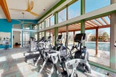 Located in Lakewood, Colorado, this fitness center offers a comprehensive and thoughtfully designed space for exercise and wellness. The main exercise room is defined by soaring vaulted ceilings, dramatic floor-to-ceiling windows framed in bold green, and vibrant multi-colored carpet tiles that lend the space a modern, energetic atmosphere. Ceiling fans circulate air throughout the generously sized room, which is bathed in abundant natural light and offers outdoor views of a neighboring pool and pergola area. The cardio zone is equipped with a full lineup of Cybex machines, including ellipticals, treadmills, and a rowing machine, while a cable strength machine and adjustable bench anchor the strength training section. Exposed ductwork along the yellow accent wall adds an industrial design touch. A dedicated studio room features warm bamboo hardwood flooring, a wall-mounted TV, spin bikes, a dumbbell rack, and a separate area with open floor space ideal for stretching or group workouts. Large windows and a green accent wall give the studio an airy, motivating feel. The facility is completed by a clean, ADA-accessible bathroom outfitted with white subway wall tile, grey floor tile, and stainless steel safety grab bars.