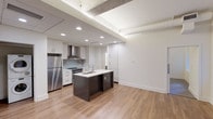 This thoughtfully designed single-floor space combines industrial character with modern finishes to create a clean, contemporary living environment. The open-concept kitchen serves as the heart of the unit, anchored by a dark-toned center island with an undermount sink and white quartz countertop. White flat-panel cabinetry lines the walls, complemented by a subway tile backsplash, a stainless steel range with a wall-mounted hood vent, and a full-size stainless steel refrigerator. Recessed lighting illuminates the space, while exposed ductwork and structural concrete elements on the ceiling lend an urban, loft-inspired aesthetic. Warm hardwood-style flooring flows seamlessly throughout, connecting the kitchen to the open living area. The living space benefits from natural light through multiple windows fitted with roller shades, offering views of the surrounding brick exterior architecture. Exposed ceiling beams add to the industrial character of the room. The bathroom is finished with light-toned large-format floor tiles, a dark vanity with a white countertop and undermount sink, an oversized mirror, and a soaking tub with a tiled surround, creating a calm and functional retreat.