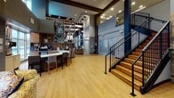 Located at 870 Toftrees Ave in State College, Pennsylvania, this single-floor community lobby space offers a welcoming and thoughtfully designed environment that blends comfort with contemporary style. The open-plan layout is anchored by warm hardwood-style flooring that flows seamlessly throughout, lending cohesion to the various functional zones within the space.

At the heart of the lobby stands a curved bar-height counter with seating, complemented by a stylish open shelving unit stocked with décor and accessories — an ideal spot for casual gatherings or quiet work. Adjacent to this, a generously sized sectional sofa in a bold geometric pattern invites relaxation, positioned to take full advantage of the expansive floor-to-ceiling windows that bathe the space in natural light and offer views of the surrounding wooded landscape.

The architecture features dramatic double-height ceilings with exposed dark wood beams and bold structural columns clad in a deep navy mosaic tile, adding a distinctive visual element. A staircase with warm wood treads and sleek black iron railings anchors one end of the space, leading to an upper landing and adding verticality to the design.

Decorative touches — including a large Earth mural, neon signage, and curated accent chairs — give the space a lively, community-oriented character that is both functional and inviting.