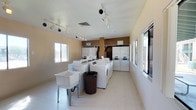 This shared laundry facility is a practical, single-floor space designed to accommodate a high volume of laundry needs efficiently. The room is furnished with a generous array of top-loading washing machines arranged in a central island configuration, complemented by stacked dryer units lining the back wall — providing substantial capacity for simultaneous use. Track lighting runs along the white ceiling, delivering bright, even illumination throughout the space and supplementing the abundant natural light that flows in through multiple windows along the walls. The windows also offer views of the surrounding outdoor greenery, lending the room an open, airy feel. Light-toned vinyl tile flooring provides a clean, durable surface underfoot. A freestanding utility sink is positioned near the entrance area, ideal for pre-treating garments or handwashing delicate items. A built-in folding counter offers a convenient surface for sorting and folding laundry. The warm earth-tone accent walls add a touch of color to the otherwise neutral interior. Two exterior doors — including a charming cross-buck style door — ensure easy access and ventilation. A fire extinguisher is visibly mounted on the wall, reflecting attention to safety. Overall, the space is functional, well-lit, and thoughtfully laid out.