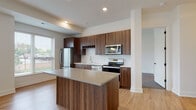 This well-appointed single-floor residence showcases a contemporary design with a thoughtful layout and quality finishes throughout. The heart of the home is an open-concept kitchen and living area, where light hardwood flooring extends across a generously sized, sun-filled space. Large windows fitted with horizontal blinds allow abundant natural light to fill the room, creating a bright and welcoming atmosphere. A modern pendant light fixture anchors the dining area, which flows seamlessly into the main living space.

The kitchen is a standout feature, equipped with rich walnut-toned cabinetry extending to the ceiling, a built-in wine rack, and a herringbone-patterned tile backsplash. Stainless steel appliances — including a refrigerator, range, over-the-range microwave, and dishwasher — complement the sleek quartz countertops. A central kitchen island provides additional workspace and a natural gathering point.

The bedrooms are carpeted and offer a calm, neutral palette with white walls and windows overlooking greenery outside. The bathroom includes a glass-enclosed walk-in shower with large-format tile surround, while a separate water closet provides added privacy. A spacious walk-in closet with built-in wire shelving offers practical, well-organized storage.