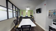 This single-floor workspace is designed as a multifunctional conference and computer room, combining collaborative meeting space with dedicated workstation areas. The room is defined by its bold green accent wall at the far end, which adds a vibrant contrast to the otherwise neutral gray walls and dark carpeted flooring. Decorative framed artwork panels are mounted against the green wall, contributing visual interest alongside a classic analog clock.

A long white central conference table is surrounded by ergonomic mesh-back chairs in black and yellow, accommodating multiple users for meetings or group work sessions. Along the green accent wall, a built-in desk runs the full width, housing several desktop computer workstations, making the space well-suited for both collaborative discussion and individual computing tasks.

A wall-mounted flat-screen television is positioned for clear visibility from the seating area. Floor-to-ceiling glass partition walls with black metal framing line one side of the room, allowing natural light to filter in while maintaining visual connectivity to the surrounding office area. Pendant lighting fixtures provide even illumination throughout. A keypad-controlled entry door with a thermostat nearby underscores the room's practical, professional design.