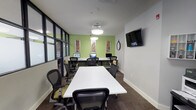 This single-floor workspace is designed as a multifunctional conference and computer room, combining collaborative meeting space with dedicated workstation areas. The room is defined by its bold green accent wall at the far end, which adds a vibrant contrast to the otherwise neutral gray walls and dark carpeted flooring. Decorative framed artwork panels are mounted against the green wall, contributing visual interest alongside a classic analog clock.

A long white central conference table is surrounded by ergonomic mesh-back chairs in black and yellow, accommodating multiple users for meetings or group work sessions. Along the green accent wall, a built-in desk runs the full width, housing several desktop computer workstations, making the space well-suited for both collaborative discussion and individual computing tasks.

A wall-mounted flat-screen television is positioned for clear visibility from the seating area. Floor-to-ceiling glass partition walls with black metal framing line one side of the room, allowing natural light to filter in while maintaining visual connectivity to the surrounding office area. Pendant lighting fixtures provide even illumination throughout. A keypad-controlled entry door with a thermostat nearby underscores the room's practical, professional design.