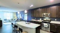 This single-floor space showcases a well-designed open-concept layout that seamlessly connects the kitchen and living area. The kitchen is a standout feature, boasting rich dark-brown cabinetry that contrasts beautifully against bold navy accent walls, creating a striking visual dynamic. A curved white quartz island takes center stage, offering bar seating for casual dining and additional prep space. The island is complemented by two modern pendant lights, adding both function and style overhead.

Stainless steel appliances, including a gas range and built-in microwave, line the kitchen wall, reinforcing a contemporary aesthetic. Ample upper cabinetry provides generous storage, while the light-toned countertops balance the darker elements throughout.

The adjacent living area flows naturally from the kitchen, maintaining the open and airy feel of the space. Light hardwood-style flooring runs throughout, enhancing the sense of continuity. The living area features natural light from a window, and the overall color palette — combining navy, white, and rich wood tones — creates a cohesive, modern ambiance that is both inviting and functional.