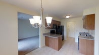 This single-floor residence presents a straightforward, functional layout designed for comfortable everyday living. The interior is finished with a neutral color palette of soft whites and warm beige tones throughout, creating a bright and cohesive atmosphere.

The kitchen serves as a central feature, equipped with medium-toned wood cabinetry, black appliances including a refrigerator, range, and microwave, and a granite-style countertop peninsula that provides additional workspace and casual seating potential. An elegant multi-light chandelier with frosted glass shades illuminates the dining area adjacent to the kitchen. Light tile flooring runs through the kitchen and entry areas, providing durability and easy maintenance.

The living area transitions seamlessly from the kitchen via an open archway and features plush carpeting underfoot. A window dressed with vertical blinds allows natural light to filter into the space. The textured ceiling adds subtle visual character throughout. Additional rooms, accessible via interior doorways, include spaces with closet storage featuring classic bi-fold doors. The overall ambiance is clean, practical, and move-in ready.