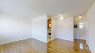 This single-floor residence offers a clean, functional layout designed for comfortable everyday living. The main living area is bright and open, showcasing polished hardwood floors that reflect natural light and lend warmth to the space. Crisp white walls provide a neutral backdrop, while a ceiling fan overhead adds both practical ventilation and a subtle decorative touch.

Adjacent to the living area, the kitchen is partially separated by a dividing wall, creating a sense of distinction between spaces while maintaining an open feel. The kitchen features warm-toned wood cabinetry that adds character and storage functionality, complemented by white appliances including a gas range and an overhead microwave. The light-colored flooring in the kitchen coordinates seamlessly with the rest of the home.

The overall layout is straightforward and efficient, making excellent use of the available floor plan. The consistent use of light tones throughout — from the walls and ceilings to the floors — creates a cohesive and airy ambiance. The space is well-suited for those seeking a tidy, move-in ready home with classic finishes and a practical design that balances openness with defined functional zones.