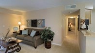 This single-floor residence offers a warm and inviting layout designed for comfortable everyday living. The living room serves as the central gathering space, featuring wall-to-wall carpeting in a soft neutral tone that adds a sense of warmth underfoot. The walls are finished in a light beige palette, creating a clean and versatile backdrop that complements a variety of décor styles. A textured ceiling adds subtle architectural character to the room.

The living area is well-proportioned, providing ample space for seating and relaxation. A hallway extends from the living room, connecting to additional rooms deeper within the residence, hinting at a functional and thoughtfully arranged floor plan. Ceiling-mounted lighting fixtures illuminate the corridor, while a secondary room visible at the end of the hall suggests comfortable private living quarters.

HVAC vents are integrated into the ceiling, ensuring consistent climate control throughout the space. The overall ambiance is calm and understated, with neutral finishes and soft lighting contributing to a relaxed, move-in-ready atmosphere suited for a range of lifestyles.