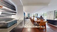 This contemporary 580 square foot space in Bethesda, MD, showcases an open-concept design that maximizes functionality and aesthetics. The single-floor layout seamlessly blends living and dining areas, creating a spacious feel despite its compact size. Floor-to-ceiling windows dominate the exterior walls, flooding the interior with natural light and offering views of the surrounding greenery.

The space features rich hardwood flooring throughout, providing warmth and continuity. The living area is furnished with a mix of modern pieces, including a plush blue sofa and elegant armchairs in neutral tones. A geometric area rug anchors the seating arrangement, adding visual interest to the space.

The dining area boasts a substantial wooden table with seating for six, perfect for entertaining or casual meals. Adjacent to this is a sleek kitchenette with dark cabinetry and state-of-the-art appliances. A striking textured accent wall behind the kitchen area adds depth and character.

Recessed lighting and strategically placed fixtures contribute to the bright, airy ambiance. The decor is minimalist yet thoughtful, with carefully chosen artwork and accessories that complement the overall modern aesthetic. This versatile space efficiently combines style and practicality, ideal for urban living or as a multifunctional area in a larger property.
