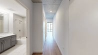 This well-appointed 2-bedroom, 2-bathroom unit showcases a contemporary urban design across a single floor. A defining architectural feature throughout the space is the exposed concrete ceiling, which adds an industrial character that contrasts beautifully with the clean white walls and warm light wood flooring found throughout.

The open-plan kitchen and living area is a standout zone, featuring sleek grey shaker-style cabinetry, quartz countertops, a white hexagonal tile backsplash, and stainless steel appliances. The living area is anchored by dramatic floor-to-ceiling windows framing expansive city views, with a structural concrete column adding to the loft-inspired aesthetic.

The primary bedroom is generously sized and benefits from large windows with frosted glass sliding panels, offering both natural light and privacy. The hallway connects the rooms with ease, leading to two well-designed bathrooms — one equipped with a soaking tub and the other with a glass-enclosed walk-in shower, both finished with large-format white tiles and chrome fixtures.

A dedicated laundry room houses a stacked Whirlpool washer and dryer unit, adding everyday convenience. The overall ambiance is modern, bright, and urban — ideal for those who appreciate refined finishes and city living.