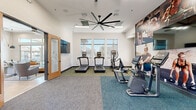 This fitness center is designed to support a full range of workout routines within a clean, modern, single-floor layout. The cardio zone anchors one side of the space, featuring multiple treadmills and elliptical machines positioned in front of a bank of large windows that allow ample natural light and outdoor views, creating an energizing environment for exercise. An oversized ceiling fan spans the center of the room, ensuring consistent airflow throughout the space.

The strength training area is equally well-appointed, offering a multi-functional cable machine and a dumbbell rack alongside an adjustable workout bench, providing options for both resistance and free-weight training. Full-length wall-to-wall motivational murals depicting athletes in action add a dynamic visual element that reinforces the fitness-focused atmosphere.

The facility is finished with durable, carpet-tiled flooring in a two-tone gray and teal scheme that delineates different workout zones. Recessed ceiling lighting and wall-mounted flat-screen televisions further enhance the user experience. A glass-paneled wooden door provides access to an adjacent area, and mirrored walls in the strength zone contribute to a sense of depth and openness. The overall design is clean, functional, and purposefully arranged to accommodate a variety of fitness activities comfortably.