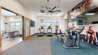 This fitness center is designed to support a full range of workout routines within a clean, modern, single-floor layout. The cardio zone anchors one side of the space, featuring multiple treadmills and elliptical machines positioned in front of a bank of large windows that allow ample natural light and outdoor views, creating an energizing environment for exercise. An oversized ceiling fan spans the center of the room, ensuring consistent airflow throughout the space.

The strength training area is equally well-appointed, offering a multi-functional cable machine and a dumbbell rack alongside an adjustable workout bench, providing options for both resistance and free-weight training. Full-length wall-to-wall motivational murals depicting athletes in action add a dynamic visual element that reinforces the fitness-focused atmosphere.

The facility is finished with durable, carpet-tiled flooring in a two-tone gray and teal scheme that delineates different workout zones. Recessed ceiling lighting and wall-mounted flat-screen televisions further enhance the user experience. A glass-paneled wooden door provides access to an adjacent area, and mirrored walls in the strength zone contribute to a sense of depth and openness. The overall design is clean, functional, and purposefully arranged to accommodate a variety of fitness activities comfortably.