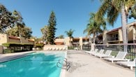 This outdoor pool area showcases a classic Spanish Colonial aesthetic, defined by warm beige stucco walls and distinctive red clay tile roofing that ties the surrounding buildings together cohesively. The generously sized swimming pool features clear, blue water and is accessible via built-in metal entry rails, making it welcoming for swimmers of all abilities.

Surrounding the pool, a spacious concrete sundeck provides ample room for relaxation, furnished with multiple white lounge chairs and outdoor seating arrangements, including umbrella-shaded tables ideal for socializing. The deck's open layout encourages a resort-like atmosphere under the abundant Southern California sunshine.

The pool area is fully enclosed by a secure wrought-iron fence with a gated entry, ensuring safety and privacy. Posted signage at the entrance clearly outlines pool hours and rules. A dedicated pool house structure with matching tile roofing provides additional functional facilities.

Mature eucalyptus, pine, and palm trees frame the entire area, creating a lush, verdant canopy that offers both shade and a sense of seclusion. The combination of Mediterranean-inspired architecture, well-maintained landscaping, and practical amenities creates an inviting and comfortable outdoor retreat.