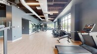 Located in Dover, NH, this single-floor fitness facility offers a well-appointed exercise environment designed to accommodate a wide range of workout styles. The main gym area features an open, airy layout with an exposed ceiling showcasing industrial-style ductwork and suspended linear LED lighting, creating a contemporary aesthetic. Light hardwood-style flooring runs throughout, providing a clean and durable surface underfoot.

The primary workout zone is generously proportioned and equipped with a comprehensive selection of cardio machines — including treadmills, ellipticals, and stationary bikes — positioned along large windows that invite abundant natural light and offer views of the surrounding greenery. A dedicated strength training area includes a multi-functional cable and rack system, kettlebells, medicine balls, resistance bands, and benches, offering a full suite of resistance training options.

Full-length mirrors line key walls throughout the space, visually expanding the room and supporting form monitoring during exercise. A separate studio room provides a more intimate setting, featuring floor-to-ceiling windows on multiple walls, recessed and LED perimeter lighting with a tray ceiling detail, and mirror-covered walls — ideal for group fitness classes, yoga, or stretching. The cool-toned blue-gray wall palette throughout lends the entire facility a calm, focused atmosphere.