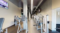 This fitness center offers a comprehensive workout environment across a single level, designed with an industrial aesthetic defined by exposed ductwork, concrete columns, and dark ceiling finishes contrasted by linear LED lighting that illuminates the space efficiently. The flooring throughout consists of durable rubber gym flooring in a speckled finish, providing both safety and comfort underfoot.

The strength training area is extensively equipped with a long row of selectorized cable and resistance machines lining the walls, complemented by a power rack with an adjustable bench and a full dumbbell rack ranging across multiple weight increments. A large mirror spans the free weights section, enhancing spatial depth and allowing users to monitor form. A small dedicated stretching and functional training alcove is visible off the main floor, outfitted with medicine balls and ab equipment.

The cardio zone features multiple treadmills and a row of elliptical cross-trainers, each positioned facing wall-mounted flat-screen televisions for entertainment during workouts. A water fountain station is conveniently located nearby. The facility also connects to an adjacent room that appears to offer additional recreational amenities. The overall ambiance is functional and well-organized, catering to a full range of fitness needs.