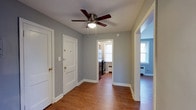 This single-floor space offers a thoughtfully connected layout centered around a main living area that serves as the heart of the home. The living space is finished with warm hardwood flooring that extends throughout, complemented by neutral gray walls and crisp white trim and baseboards that create a clean, cohesive aesthetic.

A ceiling fan with a built-in light fixture is mounted overhead, providing both air circulation and ambient lighting to the central area. Two white paneled doors are positioned along one wall — one serving as the main entry door, fitted with brass hardware, and the other providing access to an additional space.

A doorway opening leads directly into the kitchen, which is visible from the main living area. The kitchen features a refrigerator, stove, and benefits from natural light streaming through a window. An open passage on the opposite side of the living area connects to an adjacent room, also featuring hardwood floors, a window with natural light, and a baseboard radiator, suggesting a bedroom or secondary living space. The overall ambiance is bright, functional, and move-in ready.