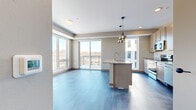 This thoughtfully designed one-bedroom, one-bathroom residence offers an efficient and modern living experience across a single floor. The open-concept kitchen and living area serves as the heart of the home, featuring rich blue-toned hardwood flooring, generous natural light from large floor-to-ceiling windows, and direct access to a private balcony with outdoor views. The kitchen is well-appointed with light wood cabinetry accented by gold hardware, a full suite of stainless steel appliances, a white subway tile backsplash, stone-topped island with an integrated sink, and elegant pendant lighting. The living area flows seamlessly from the kitchen, creating a bright and airy entertaining space. The bedroom is a comfortable retreat finished with soft carpeting, a large closet with sliding doors, and ample natural light from well-positioned windows. A hallway connects the main living spaces, with wood-look flooring continuing through the corridor. The bathroom features clean white walls, wood-look tile flooring, a soaking tub, and warm gold fixtures throughout, including a matching towel bar and tub filler, adding a cohesive, upscale touch to the overall design.