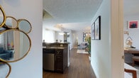 This single-floor residence offers a thoughtfully designed open-concept layout that seamlessly connects the kitchen and living areas. The kitchen features dark espresso cabinetry paired with granite-style countertops, a stainless steel dishwasher, and a peninsula that provides additional prep space and casual seating opportunities. A pendant light fixture hangs above, adding a warm, focused glow to the workspace. Rich, dark hardwood-style flooring runs throughout the main living areas, creating a cohesive and elegant foundation. The living area is bright and inviting, benefiting from natural light that filters through large windows dressed with light curtains. The textured ceiling adds subtle visual depth to the space. A neutral color palette of soft whites and grays lends the home a clean, modern aesthetic, complemented by tasteful wall art and decorative greenery. A hallway visible from the main living area suggests additional rooms beyond, contributing to the overall sense of a well-organized, functional floor plan. The combination of modern finishes, open sightlines, and warm natural tones creates a comfortable and stylish living environment throughout.
