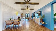 The Pointe Grand Apartment Homes leasing office and clubhouse is a thoughtfully designed single-story space that blends coastal charm with modern sophistication. The main entry welcomes visitors with soaring vaulted ceilings accented by bold dark wood beam detailing and an eye-catching geometric ceiling fixture. Underfoot, rich herringbone-patterned hardwood flooring flows throughout, tying the entire space together.

The reception area features a sleek white leasing desk adorned with the Pointe Grand logo, set against shiplap-paneled white walls that lend a clean, airy aesthetic. A dedicated seating nook with a round wood table and woven rattan chairs sits beneath a statement gold chandelier, providing a welcoming space for resident consultations. Glass double doors at the rear offer a direct view to the outdoor pool area, filling the interior with natural light.

The lounge area is anchored by a striking blue shiplap accent wall housing a wall-mounted television, complemented by plush neutral sofas and armchairs arranged around a wood coffee table. Adjacent to the lounge is a well-appointed kitchenette featuring white shaker cabinetry, dark countertops, stainless steel appliances, a beverage cooler, and a vibrant blue vertical tile backsplash with open wood shelving — perfect for a coffee and refreshment station. Together, these areas create an inviting, resort-style community hub.