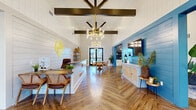 The Pointe Grand Apartment Homes leasing office and clubhouse is a thoughtfully designed single-story space that blends coastal charm with modern sophistication. The main entry welcomes visitors with soaring vaulted ceilings accented by bold dark wood beam detailing and an eye-catching geometric ceiling fixture. Underfoot, rich herringbone-patterned hardwood flooring flows throughout, tying the entire space together.

The reception area features a sleek white leasing desk adorned with the Pointe Grand logo, set against shiplap-paneled white walls that lend a clean, airy aesthetic. A dedicated seating nook with a round wood table and woven rattan chairs sits beneath a statement gold chandelier, providing a welcoming space for resident consultations. Glass double doors at the rear offer a direct view to the outdoor pool area, filling the interior with natural light.

The lounge area is anchored by a striking blue shiplap accent wall housing a wall-mounted television, complemented by plush neutral sofas and armchairs arranged around a wood coffee table. Adjacent to the lounge is a well-appointed kitchenette featuring white shaker cabinetry, dark countertops, stainless steel appliances, a beverage cooler, and a vibrant blue vertical tile backsplash with open wood shelving — perfect for a coffee and refreshment station. Together, these areas create an inviting, resort-style community hub.