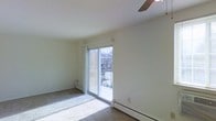 This single-floor space offers a clean, minimalist layout filled with natural light. The main living area is highlighted by a large sliding glass door that opens to a private balcony, providing views of the surrounding neighborhood and allowing abundant sunlight to flood the interior. The sliding door is framed by a simple roller shade, offering flexible light and privacy control.

The room features neutral, light-toned walls and flooring, creating a bright and open atmosphere. A ceiling fan is installed overhead, contributing to year-round comfort and air circulation. Baseboard heating units run along the lower portions of the walls, ensuring efficient warmth throughout the colder months.

The overall layout is open and uncluttered, making the space highly adaptable for a variety of furniture arrangements and uses. The combination of the generous sliding door, the ceiling fan, and the baseboard heating system reflects a practical and functional design approach. The connection to the outdoor balcony further enhances the livability of the space, extending the usable area beyond the interior and offering a pleasant outdoor retreat. Overall, this space presents a straightforward, well-lit environment suited for comfortable everyday living.