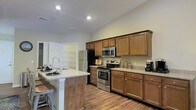 This single-story home offers a thoughtfully designed layout centered around a standout kitchen. The kitchen is a functional and stylish hub, featuring warm, rich-toned wood cabinetry with brushed nickel hardware that provides ample storage. Granite-style countertops extend across the workspace and onto a central island equipped with a deep sink, offering additional prep space and casual seating. Stainless steel appliances, including a gas range with oven, over-the-range microwave, and a full-size refrigerator, lend a sleek, contemporary feel. Hardwood-style flooring flows seamlessly throughout, adding warmth and continuity to the space. Recessed lighting illuminates the kitchen evenly, creating a bright and welcoming atmosphere. A walk-in pantry or utility closet is visible just off the kitchen, adding valuable storage and convenience. The light-toned walls and clean lines contribute to an open, airy ambiance. Overall, the space combines practical functionality with a comfortable, modern aesthetic, making it well-suited for both everyday living and entertaining.
