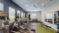 Located at 206 Cedar Branch Lane in Wilmington, NC, this single-floor fitness center is thoughtfully designed to support a comprehensive workout experience. The space features a striking navy shiplap accent wall that anchors the room and adds a bold, modern aesthetic against the light gray walls and recessed lighting overhead. Ceiling fans help maintain airflow throughout the facility.

The cardio zone is well-stocked with multiple treadmills and elliptical machines, all positioned along the accent wall and facing windows fitted with blinds that bring in natural light while maintaining a comfortable atmosphere. A dedicated strength training area includes a dual cable pulley machine, a full rack of dumbbells, and a weight bench, providing ample options for resistance training.

A vibrant green turf section defines a functional training zone, complemented by wall-mounted shelving displaying resistance bands, foam rollers, and accessories. A rack of medicine balls and wall mirrors further enhance this area, supporting dynamic and varied workouts. The rubber flooring throughout ensures safety and durability.

A glass-panel entry door connects the fitness center to an adjoining space, adding a sense of openness. Overall, the facility is clean, organized, and purposefully laid out to serve a wide range of fitness needs.