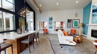 This single-floor space makes a strong design statement through its combination of high ceilings, contemporary furnishings, and a bold, curated color palette. The open layout flows seamlessly between a counter-height work or gathering area on one side and a comfortable lounge seating arrangement on the other. A geometric pendant chandelier with an industrial-modern aesthetic anchors the left side of the space, drawing the eye upward toward the dramatic ceiling height. The flooring features a distinctive terrazzo-style surface with organic patterning, adding visual texture and grounding the space. A large, white sofa accented with vivid orange throw pillows faces a sleek white coffee table, while matching orange accent chairs are positioned throughout, creating a cohesive and energetic palette. Curated wall art in complementary tones of blue, orange, and black adds personality to the crisp white walls. Interior windows allow light to pass between areas, enhancing the sense of openness. The overall ambiance is modern and design-forward, balancing functionality with an inviting, gallery-like aesthetic that encourages both productivity and relaxation.
