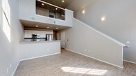 This two-story space is defined by its dramatic vertical scale, with soaring ceilings on the main level that create an open, airy atmosphere. The living area features light wood-look flooring that extends throughout, complemented by neutral taupe walls and recessed lighting. A staircase with a distinctive metal-railed upper balcony connects the two floors, adding a striking architectural element visible from the main living space.

The kitchen is efficiently designed with white cabinetry, granite-style countertops, and stainless steel appliances, including a refrigerator, range, and built-in microwave. A peninsula provides additional workspace and casual seating potential. Large windows on the main level allow abundant natural light to pour across the open floor plan.

Upstairs, the upper level features plush carpeting throughout the bedrooms, each with vaulted ceilings that follow the roofline. One bedroom includes a ceiling fan and a window with natural light. The bathrooms feature white cabinetry with granite-style countertops, wood-look flooring, and brushed nickel fixtures. The upper level also includes a versatile loft-style area with angled ceilings, offering flexible additional living or storage space.