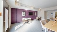 Located at 13415 Old Statesville Rd in Huntersville, NC, this single-floor community space offers a well-appointed layout comprising a dining room and a living room, thoughtfully designed for both function and comfort. The living area features a striking bold-purple wainscoted accent wall adorned with botanical artwork and a wall-mounted flat-screen television, complemented by light carpeting and recessed ceiling lighting. Flexible seating arrangements with woven-back chairs and clean-lined tables make the space ideal for gatherings or casual use. The dining area transitions seamlessly into a well-equipped utility zone featuring floor-to-ceiling natural wood cabinetry, a built-in sink, wall-mounted wicker storage baskets, and hanging aprons — suggesting dedicated space for arts, crafts, or instructional activities. Generous natural light filters in through glass-panel exterior doors that provide direct access to an outdoor deck, enhancing the sense of openness throughout. Additional amenities include a workstation nook with a desktop computer, glass-paneled French doors connecting interior areas, and recessed lighting throughout that keeps the space bright and welcoming. The overall design balances practicality with an inviting, polished aesthetic suited for community or activity-focused programming.
