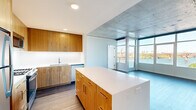 This contemporary 1-bedroom, 2-bathroom residence offers a sophisticated urban living experience defined by industrial-modern design elements and abundant natural light. Exposed concrete ceilings and walls provide a striking architectural contrast against crisp white interiors throughout the space, while wide-plank hardwood flooring anchors the open-concept kitchen and living area.

The kitchen is well-appointed with warm wood cabinetry, white quartz countertops, stainless steel appliances including a gas range, refrigerator, and dishwasher, and a generous central island that doubles as a breakfast bar. The living area opens directly to a wall of floor-to-ceiling windows and a glass door leading to a balcony, framing expansive views of the waterfront, river bridges, and city skyline.

The bedroom is generously sized and features the same dramatic floor-to-ceiling windows with unobstructed river views, exposed concrete accents, and plush carpeting. It is complemented by a spacious walk-in closet with built-in shelving and hanging rods. Both bathrooms are finished with clean, modern fixtures — one featuring a soaking tub with subway tile surround, the other a glass-enclosed shower — both with wood vanities and white quartz countertops. The overall ambiance is bright, airy, and distinctly contemporary.