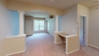 This single-floor residence offers a thoughtfully designed layout that balances open living with functional room separation. The living area serves as the heart of the space, highlighted by a graceful arched entryway flanked by decorative white columns that add architectural character. Soft, neutral-toned carpet runs throughout the living and sleeping areas, while a built-in desk or counter nook provides a convenient workspace. Large windows with vertical blinds allow natural light to fill the room, creating a bright and welcoming atmosphere. A front entryway with a white paneled door completes the living space.

The kitchen is well-appointed with warm honey-toned wood cabinetry, light countertops, and a full suite of white appliances including a refrigerator, electric range, microwave, and dishwasher. A central kitchen island offers additional prep space and casual dining potential. Track lighting overhead ensures the kitchen remains well-illuminated. Tile flooring in this area provides a practical contrast to the carpeted living zones.

The bedroom is a calm, carpeted room with a large window that brings in ample natural light. The bathroom features tile flooring, a warm wood vanity with a white countertop, and a walk-in shower stall equipped with grab bars, reflecting an accessible and safety-conscious design. Overall, the space presents a clean, neutral palette throughout.