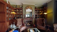 The Library Bar is a richly appointed single-floor venue that masterfully blends the warmth of a classic gentlemen's library with the character of an eclectic cocktail bar. The main lounge area anchors the space with a wood-burning fireplace framed by an ornate gilded mirror and flanked by floor-to-ceiling built-in bookshelves finished in warm wood tones. Olive-green walls, crown molding, and dark hardwood floors contribute to the refined, intimate atmosphere. A pair of tufted brown leather wingback chairs sit fireside, while an assortment of additional seating—including a vibrant teal tufted chair—adds eclectic personality throughout the room. Tall arched windows along one wall admit generous natural light and offer views of the surrounding streetscape, offset by dramatic dark curtains. A mounted zebra head serves as a bold focal point near the windows, reinforcing the venue's distinctive aesthetic. The full bar area features extensive cabinetry, mirrored backsplash, dark stone countertops, and a rustic wooden bar top with quilted leather barstools. A brass drinks trolley adds a classic touch. The restroom continues the zebra theme with bold black-and-gold patterned wallpaper, dark wainscoting, and marble-effect flooring, creating a dramatic finishing detail.