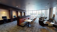 This business center is a thoughtfully designed, single-floor workspace that blends productivity and comfort seamlessly. The space is anchored by a row of private computer workstations tucked into dark-wood partitioned alcoves, each equipped with desktop computers and task lighting, offering a focused environment for individual work. Central to the layout are banquette-style seating booths with bold red upholstery and white-top tables, providing comfortable spots for collaborative work or casual meetings. Complementing these are multiple seating clusters featuring contemporary lounge chairs in neutral and dark tones arranged around low tables, ideal for informal gatherings. Floor-to-ceiling windows along one wall flood the space with natural light and offer open views of the surrounding exterior courtyard and buildings. Recessed ceiling lighting and a dark, refined ceiling finish contribute to the polished, modern aesthetic throughout. A wall-mounted flat-screen television adds a media element to the lounge area. Toward the rear, a separate glass-walled conference room with its own entrance provides a private, enclosed setting for meetings, while an adjacent kitchenette or refreshment area is visible in the background. Textured carpet tiles in muted tones complete the professional yet welcoming atmosphere of this versatile space.