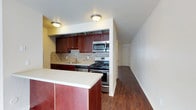 This two-floor space presents a well-appointed kitchen as a central feature, showcasing rich mahogany-toned cabinetry that extends to the upper walls, providing ample storage. The kitchen is equipped with stainless steel appliances, including a gas range, built-in microwave, and dishwasher, making it both practical and modern. A neutral-toned countertop runs along the workspace, complemented by a decorative mosaic tile backsplash in warm beige and brown tones that adds texture and visual interest. A peninsula with matching cabinetry extends into the adjoining area, offering additional counter space and defining the transition between the kitchen and the rest of the living area. Dark hardwood flooring flows seamlessly from the kitchen into the hallway beyond, adding warmth and continuity throughout the space. The walls are finished in a clean white and soft yellow palette, keeping the interior feeling bright and open. Flush-mount ceiling light fixtures provide consistent overhead illumination. A hallway visible from the kitchen suggests additional rooms beyond, reinforcing the functional, multi-room layout spread across two floors.