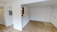 This single-floor space features a straightforward, functional layout designed to maximize connectivity between living areas. The main living area is finished with neutral-toned carpeting and bright white walls, creating a clean and versatile backdrop well-suited for a variety of furnishing arrangements. A defining architectural detail is the built-in pass-through window cut into the dividing wall, which visually and functionally connects the living space to the adjacent kitchen area. This opening allows for easy communication and light flow between the two areas while maintaining a degree of separation. The kitchen, partially visible through the pass-through, features warm wood-toned cabinetry that provides a contrast to the otherwise neutral palette of the space. The ceilings follow a standard height, and the overall layout feels open and uncluttered. The white walls throughout lend the space a bright, airy quality, even in the absence of abundant natural light sources visible in the image. The single-floor design ensures ease of movement throughout, making the space practical and accessible.