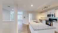Located at 3824 Banyan Grove Lane in Virginia Beach, Virginia, this single-floor, two-bedroom unit offers a clean, modern aesthetic throughout its well-designed layout. The interior is finished with light wood-look flooring in the main living areas, creating a cohesive and contemporary feel.

The kitchen is a standout feature, equipped with white shaker-style cabinetry, granite countertops, and a full suite of stainless steel appliances including a range, microwave, refrigerator, and dishwasher. A granite-topped island with an undermount sink provides additional prep space and separates the kitchen from the adjacent living areas. Track lighting illuminates both the kitchen and surrounding spaces effectively.

The living room is open and airy, benefiting from multiple windows and a sliding glass door that leads to a private outdoor balcony or patio, allowing natural light to fill the room. The bedrooms offer plush carpeting and dedicated windows.

Both bathrooms mirror the unit's design language with granite vanity tops, white cabinetry, wood-look flooring, and brushed nickel hardware. One bathroom includes a tub-shower combination with tiled surround. Throughout, the unit maintains a bright, neutral palette that lends itself to a clean and move-in-ready presentation.