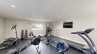 Located at 2525 Booker Creek Rd in Chapel Hill, NC, this dedicated fitness center offers a well-equipped exercise space designed to support a well-rounded workout routine. The single-room layout is finished with neutral gray walls, a textured ceiling, and recessed lighting that keeps the space bright and functional throughout the day. Dark rubber flooring covers the entire area, providing a durable, non-slip surface ideal for a variety of exercise activities.

The room is outfitted with a diverse selection of equipment, including a treadmill, elliptical trainer, stationary recumbent bike, and multiple rowing machines — offering ample options for cardiovascular training. A multi-station cable machine and an adjustable weight bench round out the strength-training section, complemented by a full dumbbell rack stocked with a range of free weights. A wall-mounted flat-screen television provides entertainment during workouts.

Multiple windows allow natural light to enter the space, creating an open and airy atmosphere despite the room's compact footprint. A wall-mounted HVAC unit ensures year-round climate comfort. Safety essentials, including a fire extinguisher and a paper towel dispenser, are also present. Overall, this fitness center provides a comprehensive and practical environment for residents seeking convenient on-site exercise facilities.