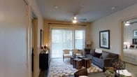 This single-floor living space offers a thoughtfully designed interior centered around a comfortable and stylish living area. The room features hardwood-style flooring that adds warmth and a sense of continuity throughout the space. A ceiling fan with an integrated light fixture is centrally positioned overhead, providing both functionality and a finishing architectural touch to the room.

The living area is filled with natural light, courtesy of a large sliding glass door dressed with vertical blinds and sheer curtains, offering both privacy and brightness control. The door provides direct access to an outdoor area, enhancing the open and airy feel of the room. Neutral-toned walls complement the overall design palette, creating a calm and inviting atmosphere.

The space is furnished with a gray sofa accented by decorative pillows, a pair of cream-colored accent chairs, and rustic wooden side tables that add a contrasting organic element. A plush, textured area rug anchors the seating arrangement, adding depth and comfort underfoot. A dark-toned console table along one wall provides functional storage and display space, completing the well-balanced and livable design of this interior.