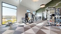 This contemporary fitness center is thoughtfully designed to accommodate a wide range of workout styles and training needs. The main gym area features soaring vaulted ceilings that create an open, airy atmosphere, enhanced by an abundance of natural light streaming through large floor-to-ceiling windows that offer views of the surrounding landscape. A bold geometric-patterned carpet in neutral tones of gray, brown, and white adds a distinctive visual element underfoot throughout this zone.

The main area is well-equipped with multiple treadmills, elliptical trainers, a stair climber, a rowing machine, and a multi-station cable strength machine. A tiered dumbbell rack provides a comprehensive range of free weights, while a punching bag station adds a combat training option. An accent wall clad in textured green paneling lends a modern, sophisticated touch.

An adjoining studio space features light wood-look flooring, full-length mirrored walls, and a wall-mounted flat-screen TV, making it ideal for group classes or individual training. This room is stocked with stability balls, medicine balls, resistance bands, and balance trainers. A dedicated equipment closet and a hydration station with a water fountain complete the thoughtfully appointed facility.