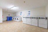 Located at 857 James St in Woodstock, Ontario, this shared laundry facility offers a practical and well-organized space designed to serve building residents efficiently. The room is finished with light-toned ceramic floor tiles throughout, contributing to a clean and easy-to-maintain environment. Bright fluorescent lighting fixtures mounted on the textured white ceiling ensure the space is well-illuminated at all times.

Along one wall, a row of front-loading washing machines is neatly aligned, complemented by a separate bank of top-loading dryers, providing ample capacity for multiple users. Instructional signage is posted above the machines to assist residents with proper operation. A stainless steel utility sink is positioned in a recessed alcove area, offering a convenient hand-washing or pre-treatment station. A baseboard heater runs along the lower wall, maintaining a comfortable temperature year-round.

The space also includes a folding table for laundry sorting, blue recycling bins for waste management, and a fire extinguisher for safety compliance. A washroom is accessible directly from the laundry room, adding to the overall convenience of the facility. The open, uncluttered layout allows for comfortable movement throughout the room, making this a highly functional shared amenity.