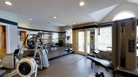 This fitness center is designed to accommodate a comprehensive workout experience within a single-level layout. The space is outfitted with a diverse selection of exercise equipment, including cardio machines such as treadmills and ellipticals, as well as strength training stations and free weights, providing users with a full range of workout options.

The flooring consists of durable rubber matting, which offers both safety and comfort during exercise. A large mirrored wall runs along one side of the room, visually expanding the space and allowing users to monitor their form while training. Recessed lighting in the white ceiling ensures the interior is well-lit throughout.

Natural light enters through large windows fitted with horizontal blinds and a set of glass-paneled doors that open to an outdoor area, enhancing the overall ambiance and providing ventilation. A decorative dark horizontal band along the upper portion of the walls adds a subtle design element. The layout is thoughtfully organized, keeping equipment accessible while maintaining open floor space for stretching or floor exercises.