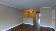 This single-floor residence offers a well-designed, open-concept layout that maximizes the sense of space and flow throughout the interior. The main living area is anchored by rich, dark hardwood flooring that runs continuously through the space, providing a warm and grounded aesthetic. Neutral gray walls complement the clean white baseboards and crown molding that frame each room, adding a polished, classic architectural detail throughout.

A defining feature of the living space is the breakfast bar peninsula, which creates a natural division between the living and kitchen areas while maintaining an open, connected feel. The bar-height counter with overhang seating is ideal for casual dining or entertaining. Overhead, a ceiling fan with an integrated light fixture provides both comfort and practical illumination.

A hallway visible from the main living area leads further into the home, hinting at additional rooms beyond. Natural light filters in from a window at the end of the corridor, brightening the interior and enhancing the airy quality of the space. The overall ambiance is clean, neutral, and move-in ready, offering a versatile canvas suited to a variety of interior styles and furnishing preferences. The thoughtful layout and consistent finishes throughout give this single-level home a cohesive and comfortable character.
