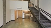 This two-story loft showcases an industrial-modern aesthetic defined by soaring ceilings and an open, airy feel. Polished concrete floors extend throughout the main level, adding a sleek, contemporary foundation to the space. The open-concept layout seamlessly connects the living area to the kitchen, which features dark wood cabinetry, stainless steel appliances including a full-size refrigerator, microwave, and range, as well as a functional sink and ample counter space. A wall-mounted split-unit air conditioner ensures climate control, while track lighting illuminates the lower level. A striking steel staircase with cable railings anchors the right side of the space, leading to the upper level mezzanine, which overlooks the main floor below through an open railing. The upper level adds versatility, suitable for use as a sleeping area, office, or lounge. The tall, exposed walls contribute to the loft's expansive character, while the combination of raw materials — concrete, steel, and wood tones — creates a cohesive industrial ambiance throughout.