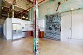 This single-floor industrial loft masterfully balances historic character with contemporary upgrades. The open-plan living and kitchen area immediately impresses with soaring ceilings that expose original steel roof framing, painted in rich reds and greens that echo the building's storied past. Weathered, paint-layered steel columns anchor the space, while polished concrete floors stretch throughout, adding warmth and continuity.

The kitchen is a standout feature, showcasing a two-tone cabinet design — rich wood lower cabinets paired with crisp white upper cabinets — complemented by granite countertops, a hexagonal tile backsplash, and a full suite of stainless steel appliances including a gas range, dishwasher, and refrigerator. Dramatic aged brick walls and an original oversized industrial sliding door serve as striking architectural backdrops in the kitchen area, lending undeniable authenticity.

A long, clean corridor connects the main living areas to the private bedroom and bathroom. The bedroom offers generous proportions, a built-in closet, and an interplay of original structural elements overhead. The bathroom is neatly appointed with warm wood cabinetry, granite countertops, a full tub-shower combination, and clean white finishes. Throughout, exposed ductwork and structural elements celebrate the building's industrial heritage while modern upgrades ensure comfort and functionality.