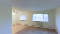This single-floor residential space offers a clean and functional layout designed for comfortable everyday living. The main living area is notably spacious, featuring light-toned walls that contribute to a bright and airy atmosphere. Multiple windows, including a large picture window, allow generous natural light to fill the room while offering views of the surrounding wooded scenery. A ceiling fan with an integrated light fixture is centrally positioned, providing both ventilation and ambient lighting. The flooring throughout the living area consists of wall-to-wall carpeting in a neutral tone, adding warmth underfoot. Climate control is well-considered, with baseboard heating running along the perimeter of the room and a wall-mounted air conditioning unit positioned beneath the large window, ensuring year-round comfort. The smooth, flat ceiling and simple trim work give the space a modest, understated character. The overall ambiance is calm and unfussy, making it a versatile canvas well-suited for a variety of furnishing styles. The abundance of natural light, combined with the open layout, creates a welcoming environment that feels open and livable throughout the day.