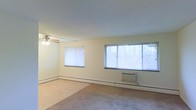 This single-floor residential space offers a clean and functional layout designed for comfortable everyday living. The main living area is notably spacious, featuring light-toned walls that contribute to a bright and airy atmosphere. Multiple windows, including a large picture window, allow generous natural light to fill the room while offering views of the surrounding wooded scenery. A ceiling fan with an integrated light fixture is centrally positioned, providing both ventilation and ambient lighting. The flooring throughout the living area consists of wall-to-wall carpeting in a neutral tone, adding warmth underfoot. Climate control is well-considered, with baseboard heating running along the perimeter of the room and a wall-mounted air conditioning unit positioned beneath the large window, ensuring year-round comfort. The smooth, flat ceiling and simple trim work give the space a modest, understated character. The overall ambiance is calm and unfussy, making it a versatile canvas well-suited for a variety of furnishing styles. The abundance of natural light, combined with the open layout, creates a welcoming environment that feels open and livable throughout the day.