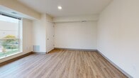 This single-floor unit presents a clean, contemporary design highlighted by warm wood-look vinyl flooring that runs consistently throughout the space, lending a cohesive and welcoming feel to every room. The kitchen is well-appointed with gray shaker-style cabinetry accented by matte black hardware, white appliances including a range, dishwasher, and microwave, and a double-basin stainless steel sink set into light-toned countertops. The kitchen flows naturally into an open living area, where a large floor-to-ceiling window fills the space with abundant natural light and provides views of the surrounding urban landscape. Recessed lighting supplements the natural light throughout. The bedroom areas feature the same neutral, cream-toned walls and consistent flooring, offering versatile, move-in-ready spaces. The bathroom is efficiently designed, finished in crisp white tones with a tub-shower combination, a toilet, and a vanity with gray cabinetry matching the kitchen's aesthetic. The overall design philosophy emphasizes simplicity and functionality, with a neutral color palette of whites, creams, and warm wood tones creating a bright, airy ambiance that provides an excellent foundation for personalization.