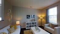 Located at 5730 Silver Falls St in Dublin, Ohio, this single-floor residence offers a thoughtfully designed living space with a clean, contemporary aesthetic. The living room serves as the heart of the home, finished in cool gray tones that lend a calm, modern atmosphere. Dark hardwood-style flooring grounds the space and contrasts elegantly with the light furnishings, including a white sofa adorned with mustard yellow and patterned accent pillows that introduce warmth and personality. A striped area rug further defines the seating area, while a white open-cube shelving unit displays decorative accents and provides practical storage. Metallic spherical décor pieces and framed wall art add visual interest throughout. Ambient lighting from a tall floor lamp and a table lamp creates a warm, inviting glow. Decorative circular mirrors arranged on one wall add depth and a sculptural quality to the room. An open pass-through connects the living area to the kitchen, which features white cabinetry, dark countertops, and stainless steel appliances, including a built-in microwave. The seamless flow between the living and kitchen areas makes the space feel open and connected, ideal for both everyday living and entertaining.