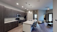 This single-floor unit showcases a contemporary design highlighted by a neutral color palette, wood-style flooring, and recessed lighting throughout. The open-concept kitchen is a standout feature, equipped with dark espresso cabinetry extending to the ceiling, granite-style countertops, and a full suite of stainless steel appliances including an electric range, built-in microwave, and refrigerator. A generously sized kitchen island with matching countertops provides an integrated sink and bar seating for multiple occupants, with pendant lighting suspended above to define the space. The kitchen flows seamlessly into the living area, which offers ample room for seating and relaxation.

The bedrooms are well-proportioned, each featuring carpet flooring, a dedicated study desk with task chair, and a window providing natural light and exterior views. Each bedroom connects directly to a private en-suite bathroom finished with granite-style countertops, dark wood cabinetry, an oversized mirror, and modern chrome fixtures. The overall design is clean, functional, and consistent in its modern aesthetic across all rooms.